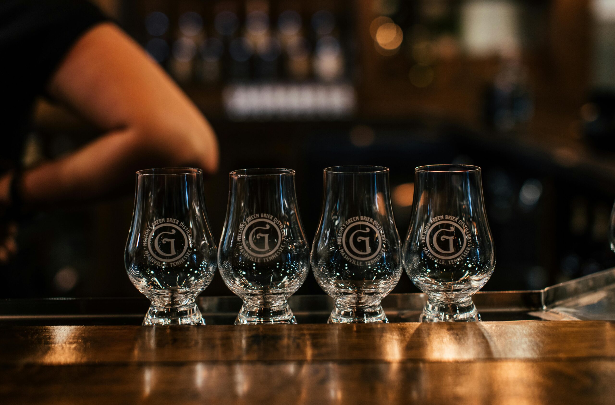 Four empty Glencairn glassesWhiskey Bottles Wholesale, Wholesale Whiskey Glasses, Wholesale Whiskey Barrels, Wholesale Whiskey, Whiskey Wholesale aligned on a bar counter in a dimly lit setting.