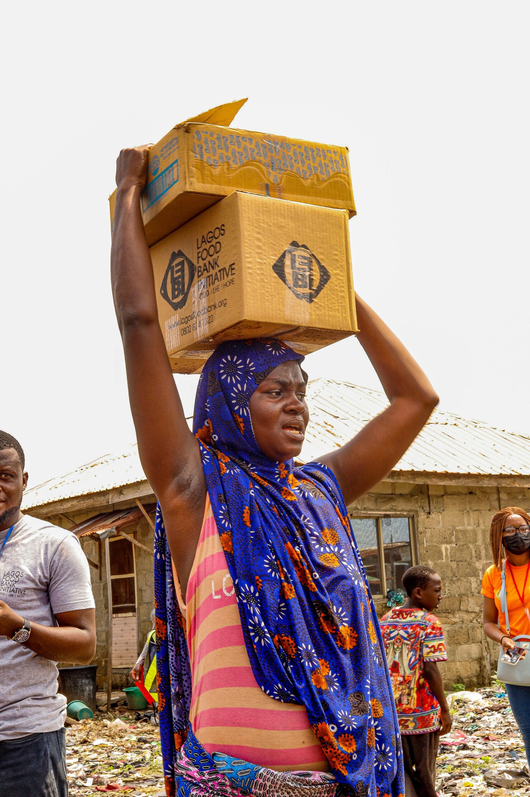 African woman in vibrant headscarf carrying boxes in a lively market scene.